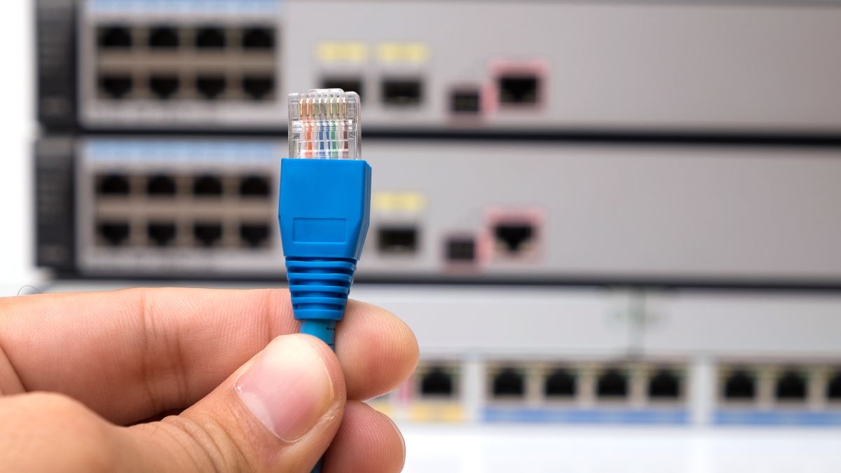 A mão de uma pessoa segurando um conector de cabo Ethernet na frente de switches de Internet.