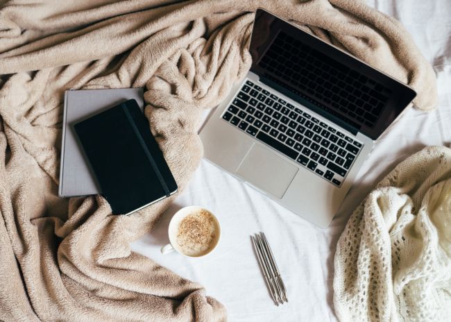 Laptop em uma cama ao lado de um diário e uma xícara de café.