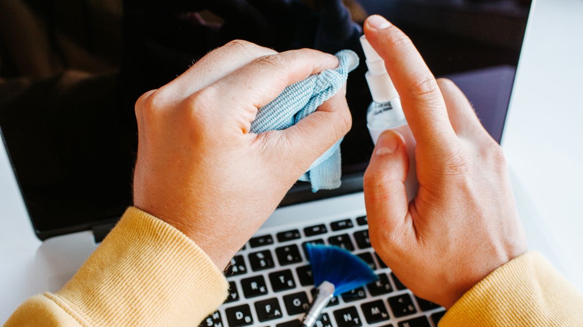 Mãos segurando um pano de microfibra e um spray de limpeza sobre um laptop aberto com uma escova sobre o teclado.