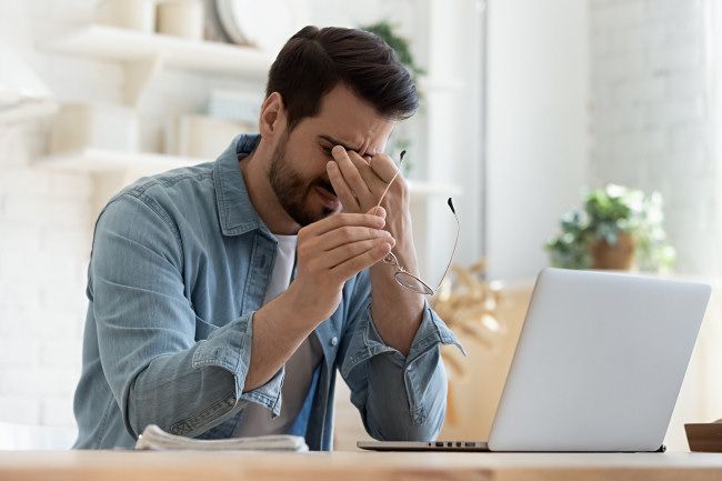 Um homem sentado em frente a um laptop com cansaço visual, esfregando os olhos.