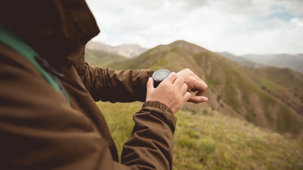As mãos de um caminhante verificando seu smartwatch, com colinas selvagens ao fundo.