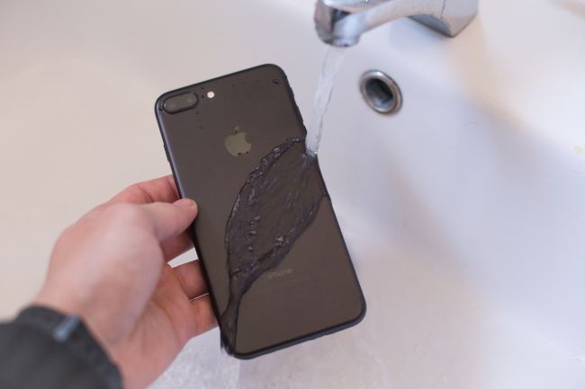 Pessoa segurando um iPhone sob água corrente de uma torneira.