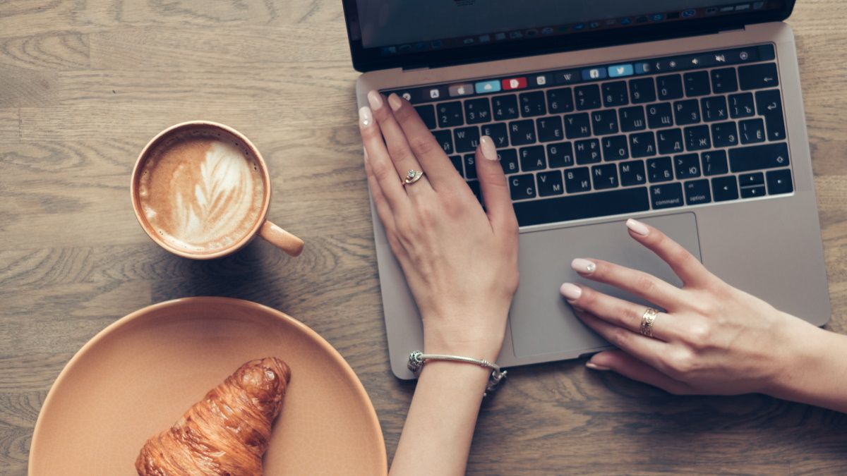 Mãos no teclado do MacBook ao lado de um café e um croissant.