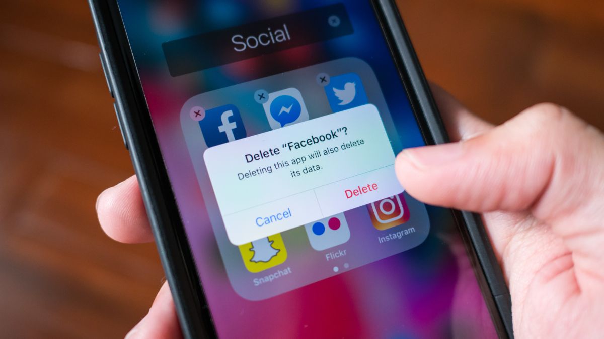 Mão de uma pessoa segurando um iPhone com uma caixa de diálogo para excluir o aplicativo do Facebook visível na tela.