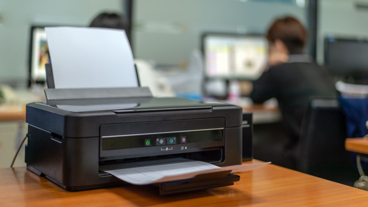Uma impressora de escritório em uma mesa com uma pessoa ao fundo usando um computador.