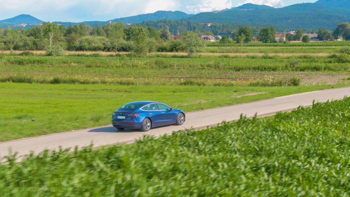 Um Tesla Model 3 azul dirigindo em uma estrada de terra através de pastos verdejantes.