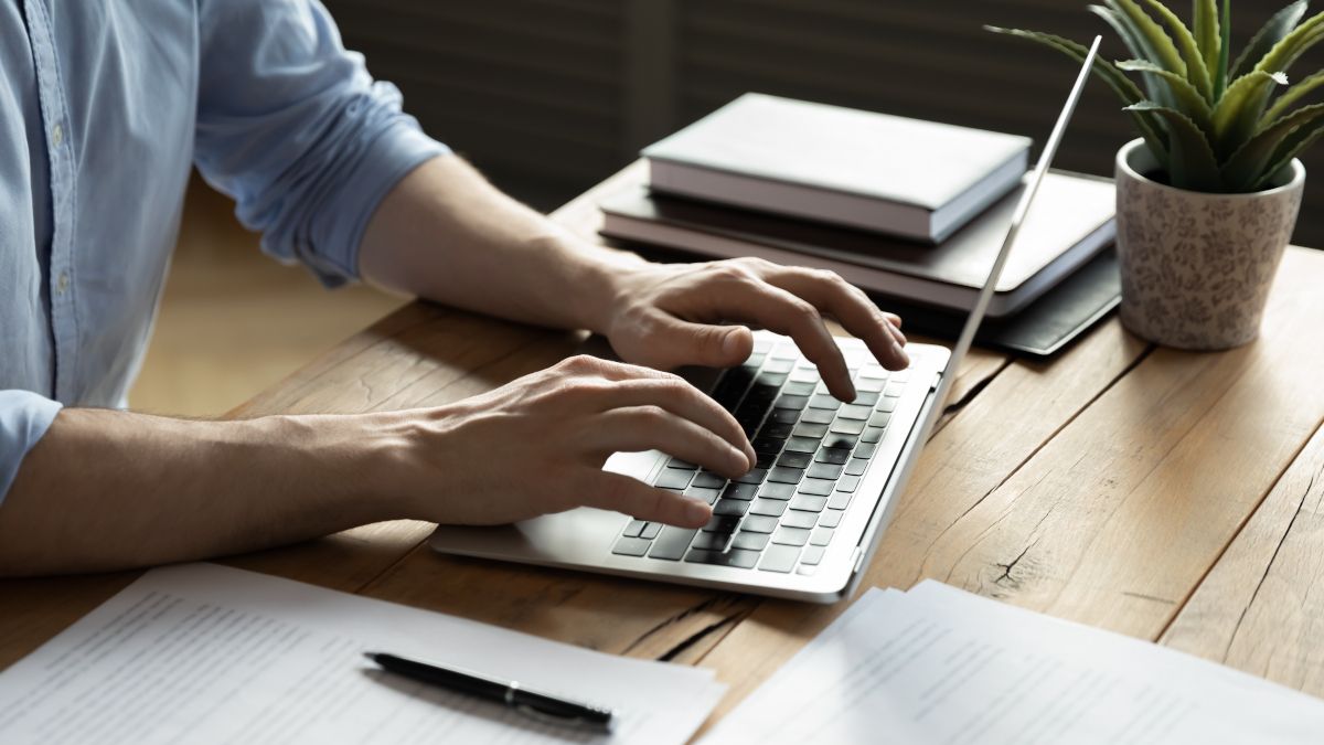 Mãos de uma pessoa digitando em um laptop com papelada e cadernos em cima da mesa.