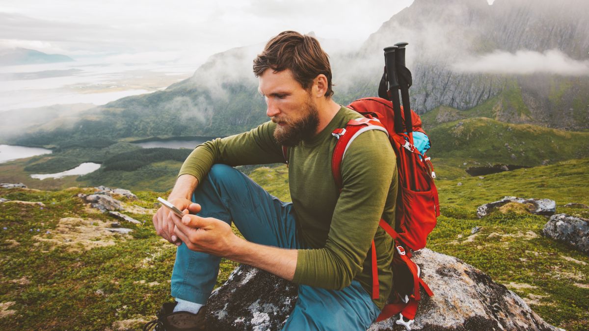Homem vestindo uma mochila e usando um smartphone no topo de uma montanha.