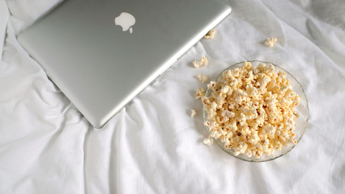 Um MacBook prateado fechado em cima de uma cama ao lado de uma tigela de pipoca.