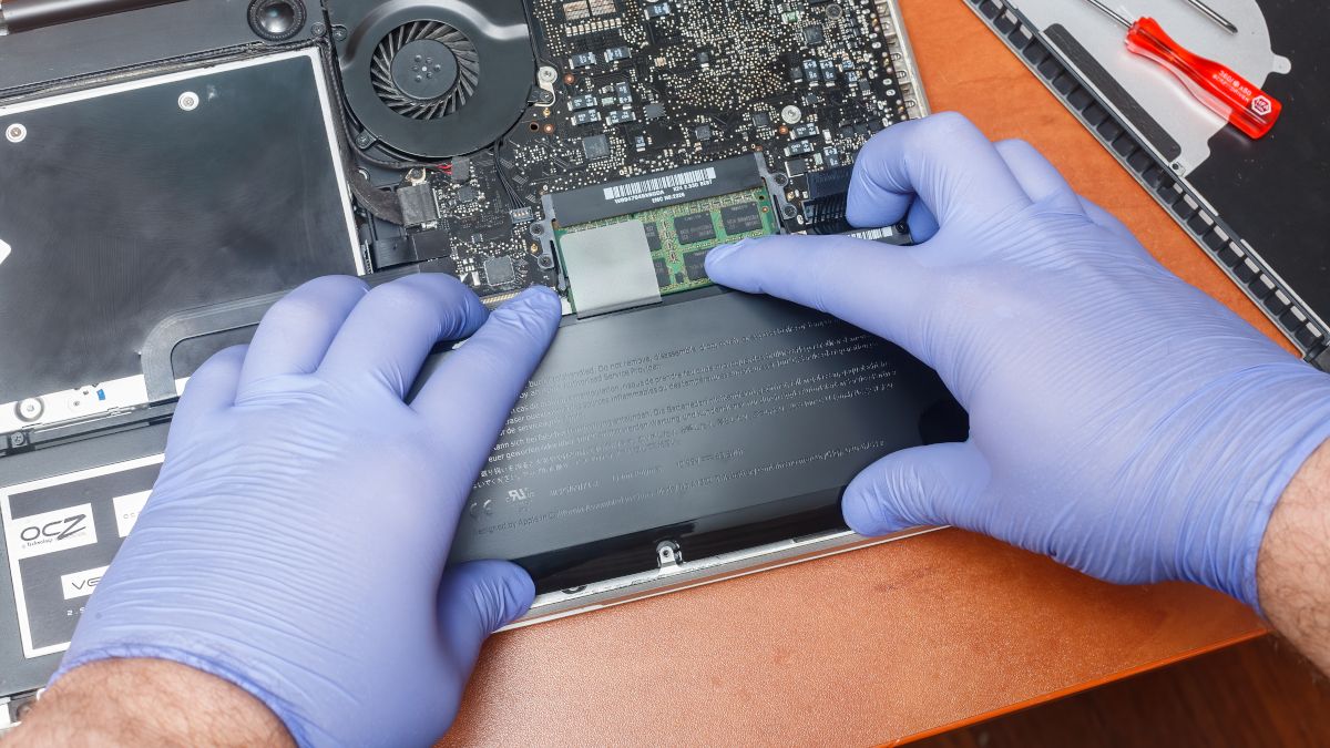 Close das mãos enluvadas de uma pessoa instalando uma nova bateria dentro de um Apple MacBook.