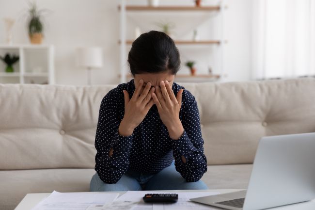 Mulher pressionando as mãos na testa, estressada, com uma calculadora e um laptop à sua frente.