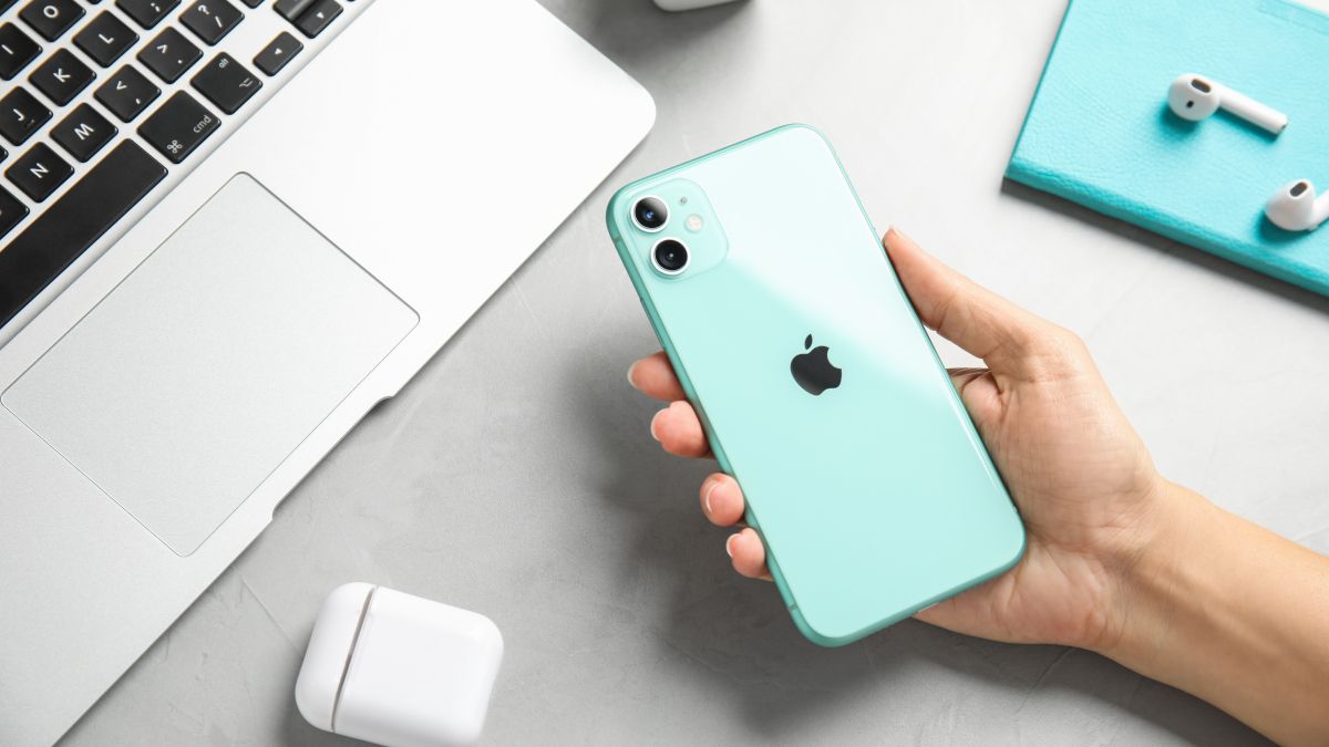 A mão de uma mulher segurando um iPhone 11 verde com AirPods e um MacBook visíveis ao fundo.