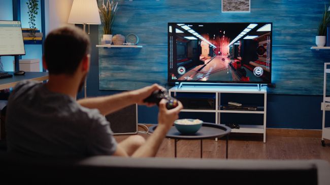 Vista por cima do ombro de um homem jogando um jogo FPS em uma TV.