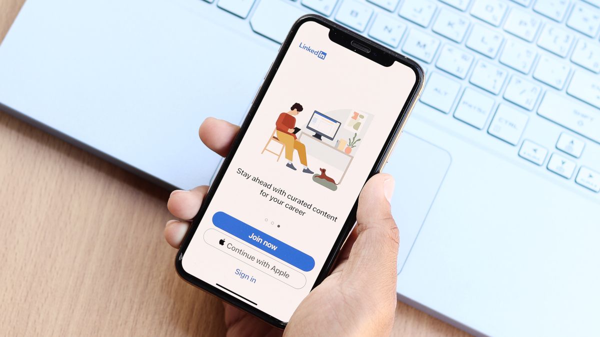 A mão de uma pessoa segurando um smartphone sobre o teclado de um laptop, com o aplicativo LinkedIn em exibição.