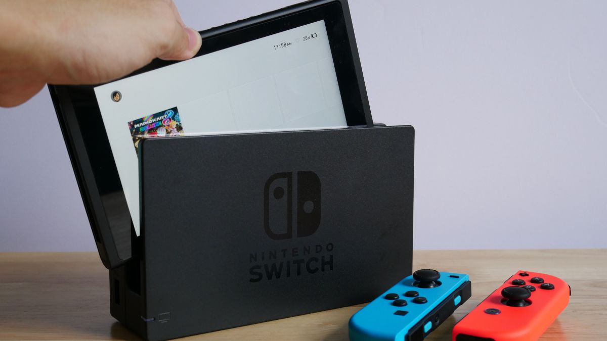 A mão de uma pessoa colocando um Nintendo Switch no dock, ao lado dos controles azuis e vermelhos.