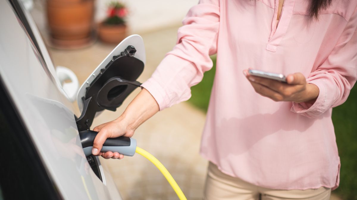 Mulher carregando um carro elétrico enquanto olha para um smartphone.