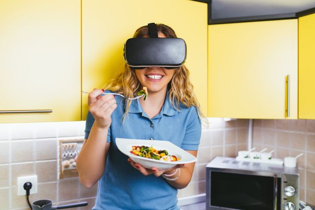 Mulher em uma cozinha usando um fone de ouvido de realidade virtual e comendo uma tigela de comida.