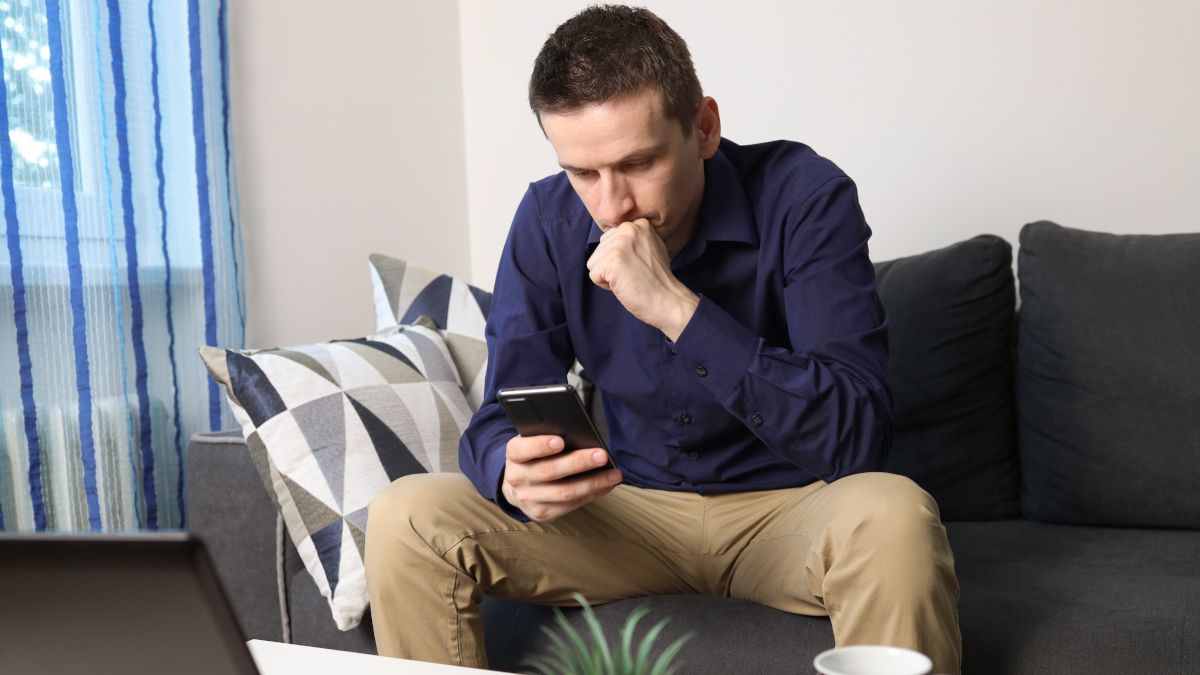 Um jovem sentado em um sofá olhando para o telefone com uma expressão preocupada.